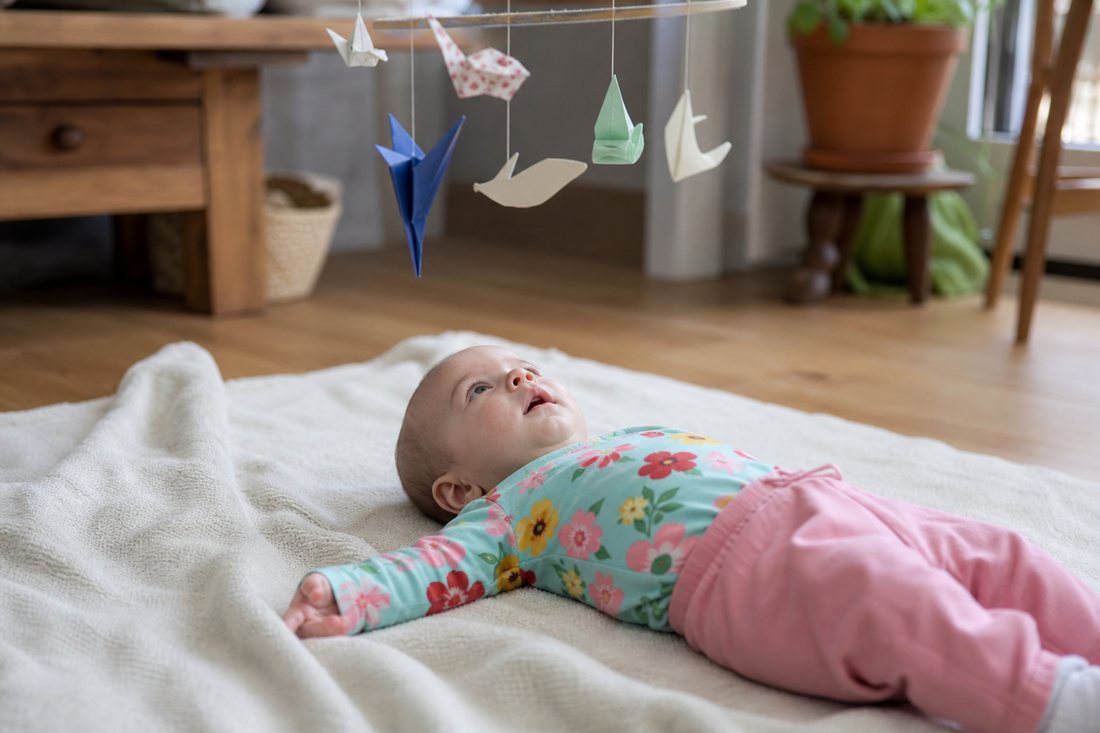 Ein Saeugling liegt auf einer Decke und betrachtet aufmerksam ein sich langsam drehendes Mobile aus Papier ueber sich.