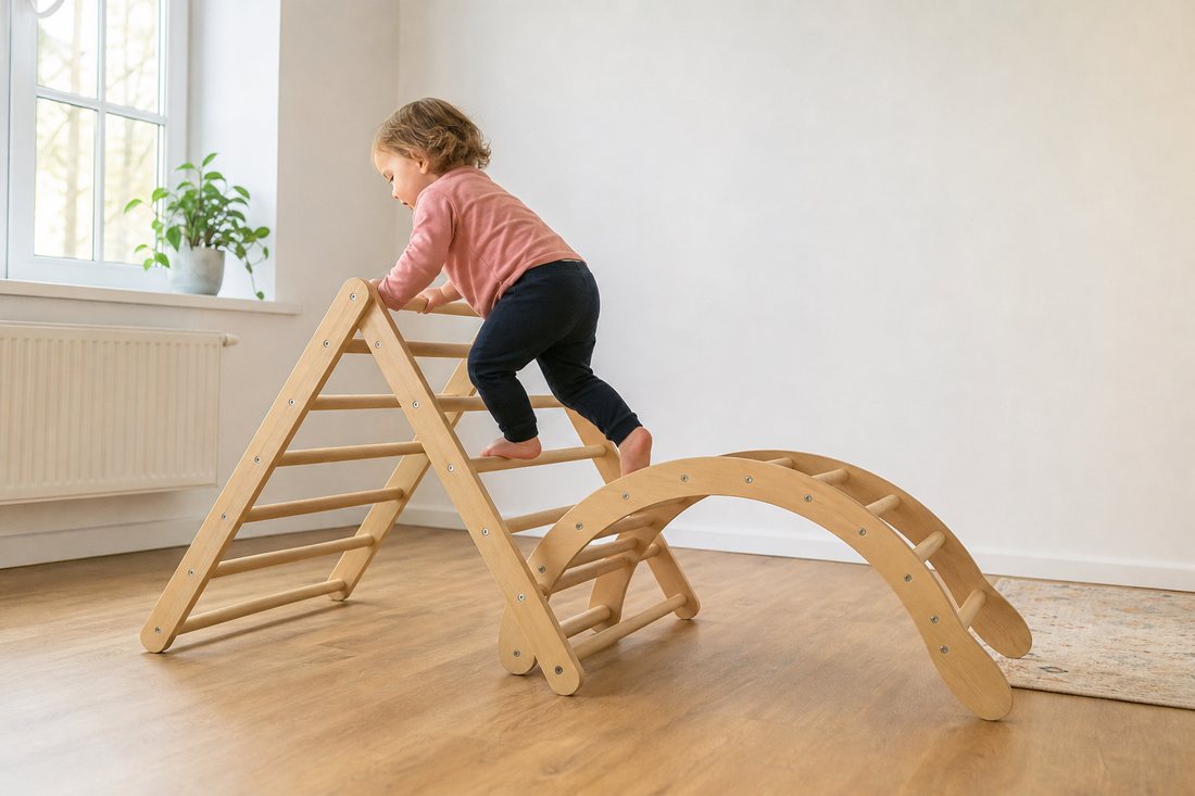 Ein Pikler-Dreieck und ein Kletterbogen aus hellem Holz in einem sonst leeren Raum, ein Kleinkind klettert langsam hinau