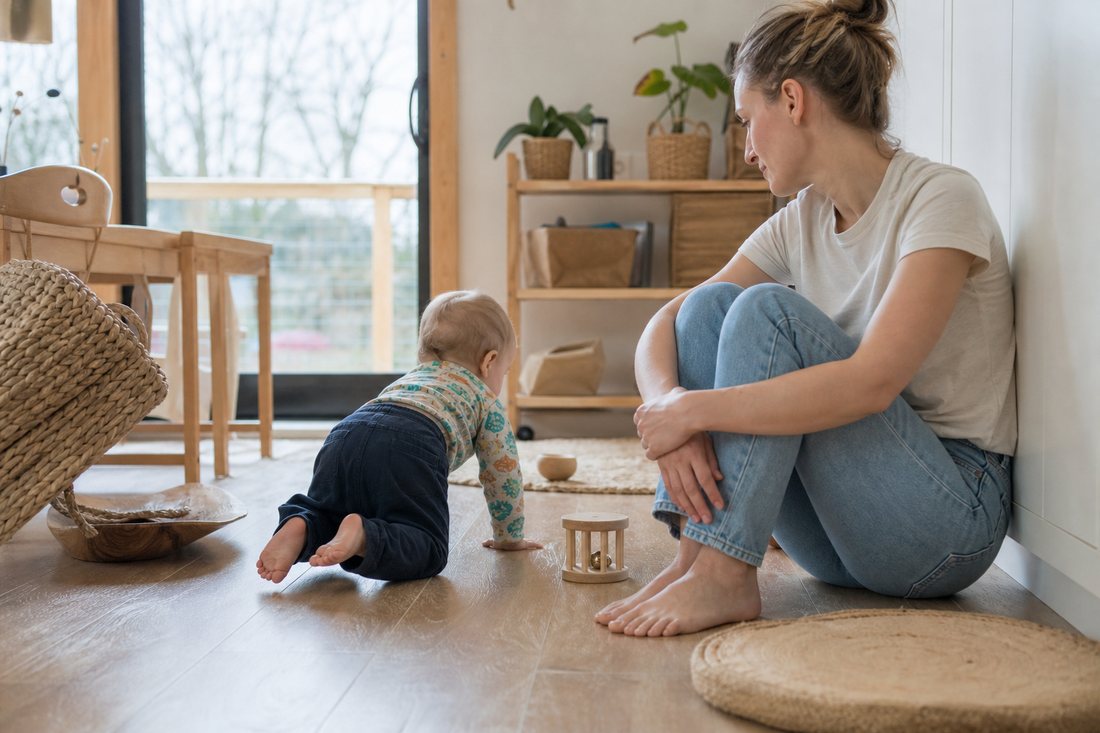 Eine erwachsene Person sitzt am Boden und beobachtet ein krabbelndes Kleinkind, ohne einzugreifen, ruhige helle Umgebung