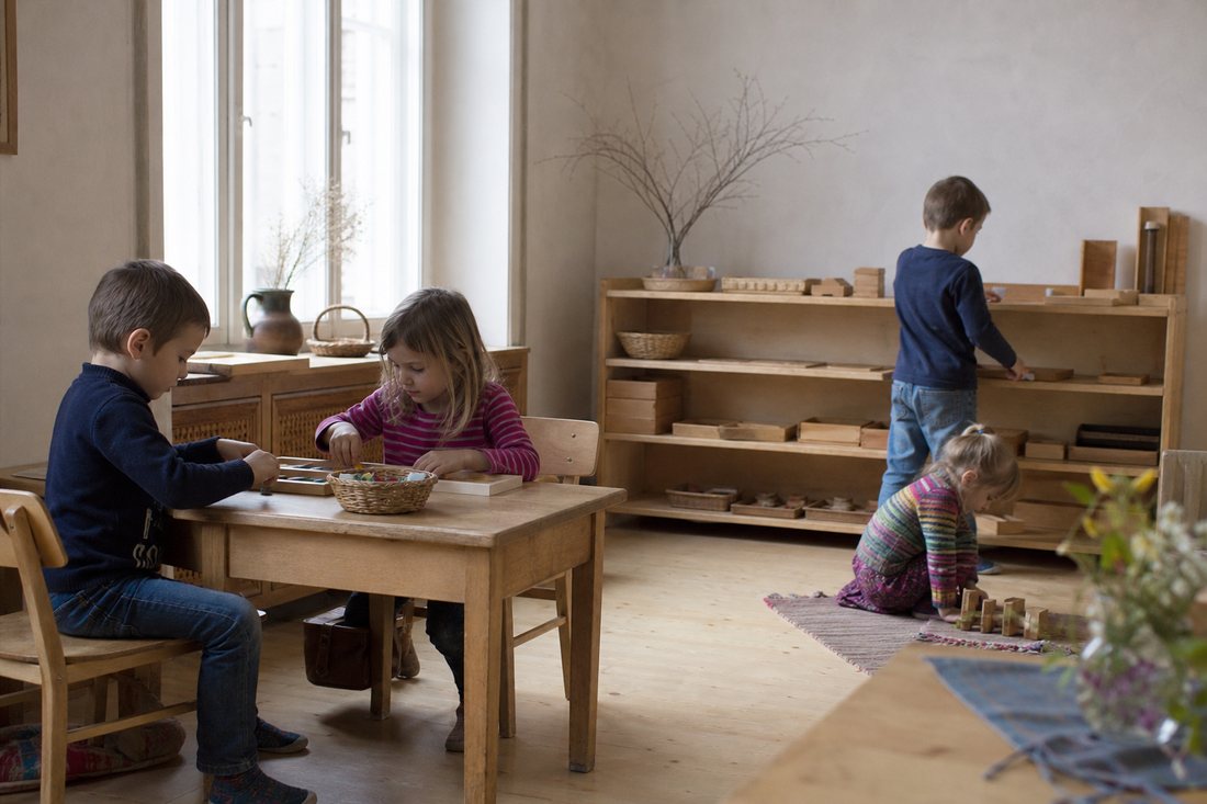 Ein einfacher Raum um 1910 mit kleinen Holztischen und Stuehlen in Kindergroesse, an der Wand ein Regal mit selbstgemach