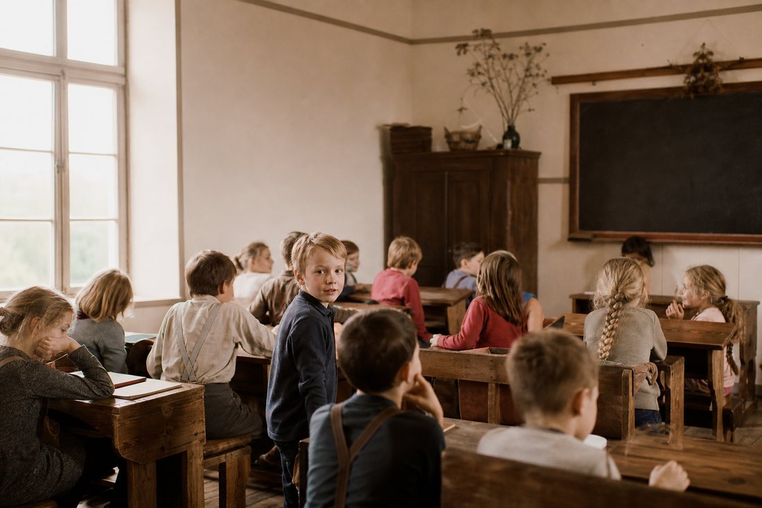 Ein historisch wirkender Klassenraum um 1910 mit einfachen Holzbaenken, Tafel und Sonnenlicht durch hohe Fenster, warme