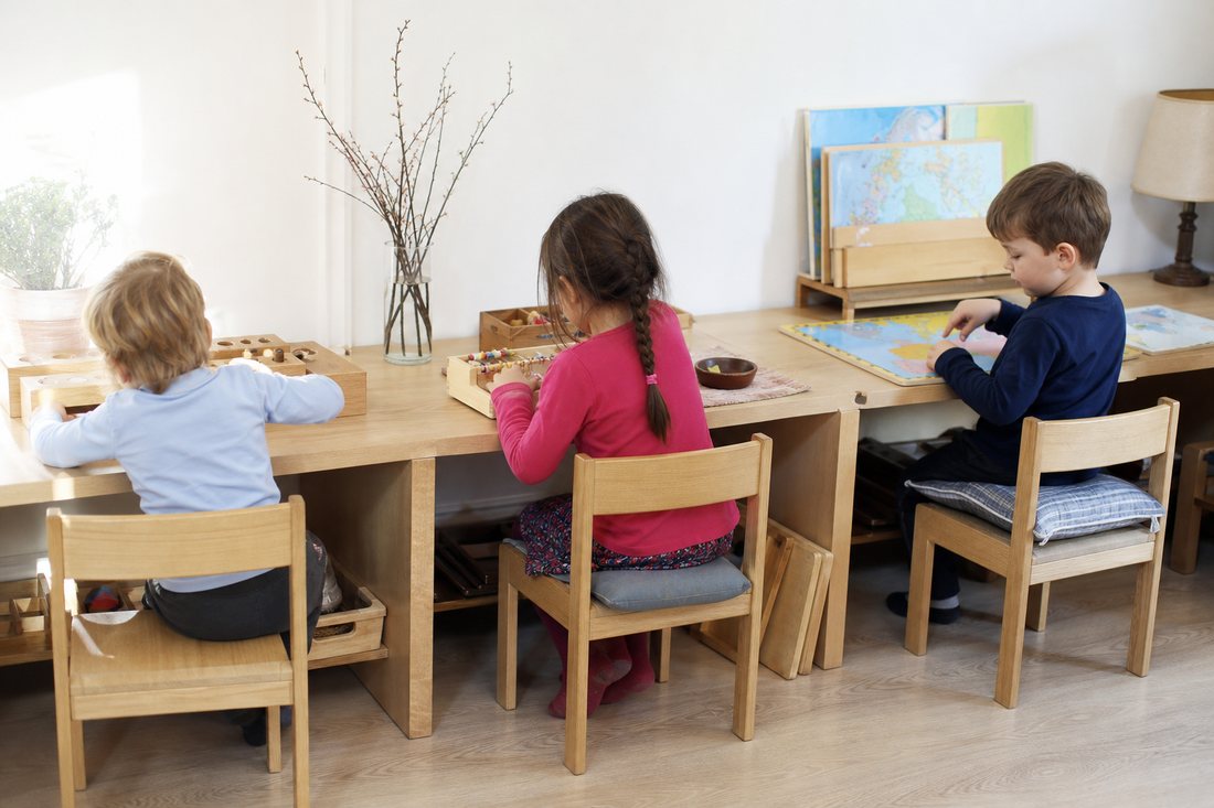 Drei Arbeitsplaetze nebeneinander in einem hellen Raum, jeder mit Material fuer ein anderes Alter: Steckspiel, Perlenmat