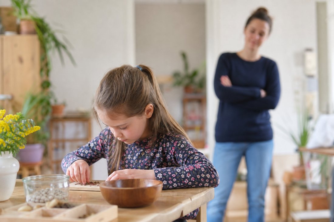 Ein Kind arbeitet konzentriert an einem kleinen Tisch, im unscharfen Hintergrund steht eine erwachsene Person und beobac