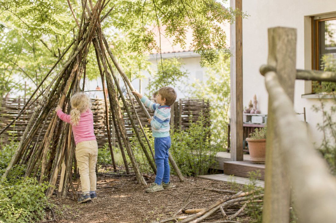 Ein einfaches Tipi aus langen Aesten und Zweigen im Garten, Kinder lehnen weitere Aeste an.