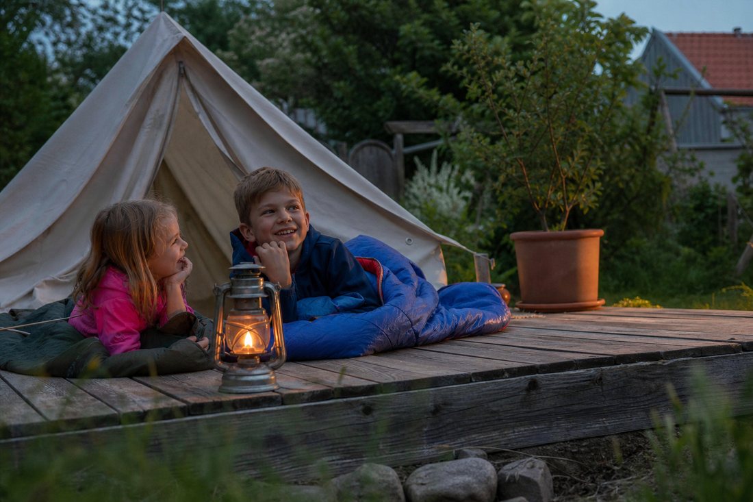 Ein kleines Zelt im Garten in der Abenddaemmerung, davor eine Laterne und zwei Schlafsaecke.