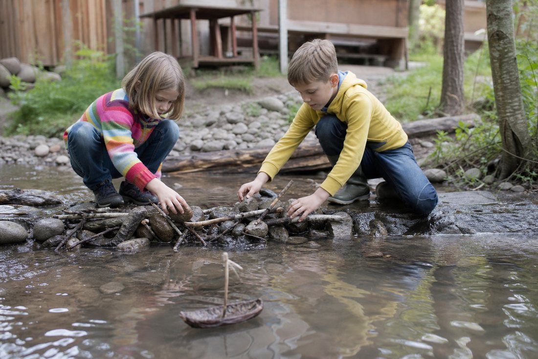 Kinder bauen an einem flachen Bach einen kleinen Damm aus Steinen und Aesten, daneben schwimmt ein Rindenschiffchen.