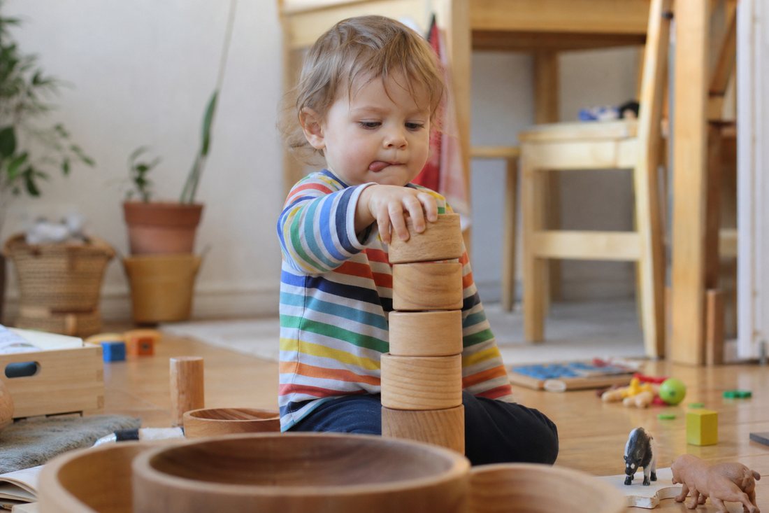Ein etwa zweijaehriges Kind stapelt hochkonzentriert Holzbecher, die Zunge im Mundwinkel, waehrend um es herum unbeachte