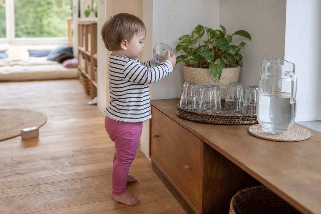 Ein Kleinkind holt sich selbst ein Glas Wasser von einer niedrigen Anrichte mit Krug und Glaesern in Kinderhoehe.