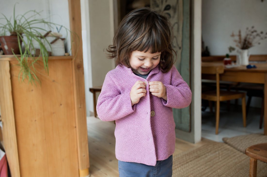 Ein Kind hat gerade seine Jacke selbst zugeknoepft und schaut mit strahlendem Gesicht an sich herunter.