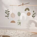 Ein schlichtes Mobile aus Holzstäben und Papierformen in gedeckten Farben hängt vor einer hellen Wand und wirft weiche