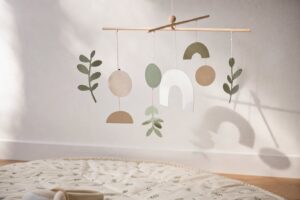 Ein schlichtes Mobile aus Holzstäben und Papierformen in gedeckten Farben hängt vor einer hellen Wand und wirft weiche
