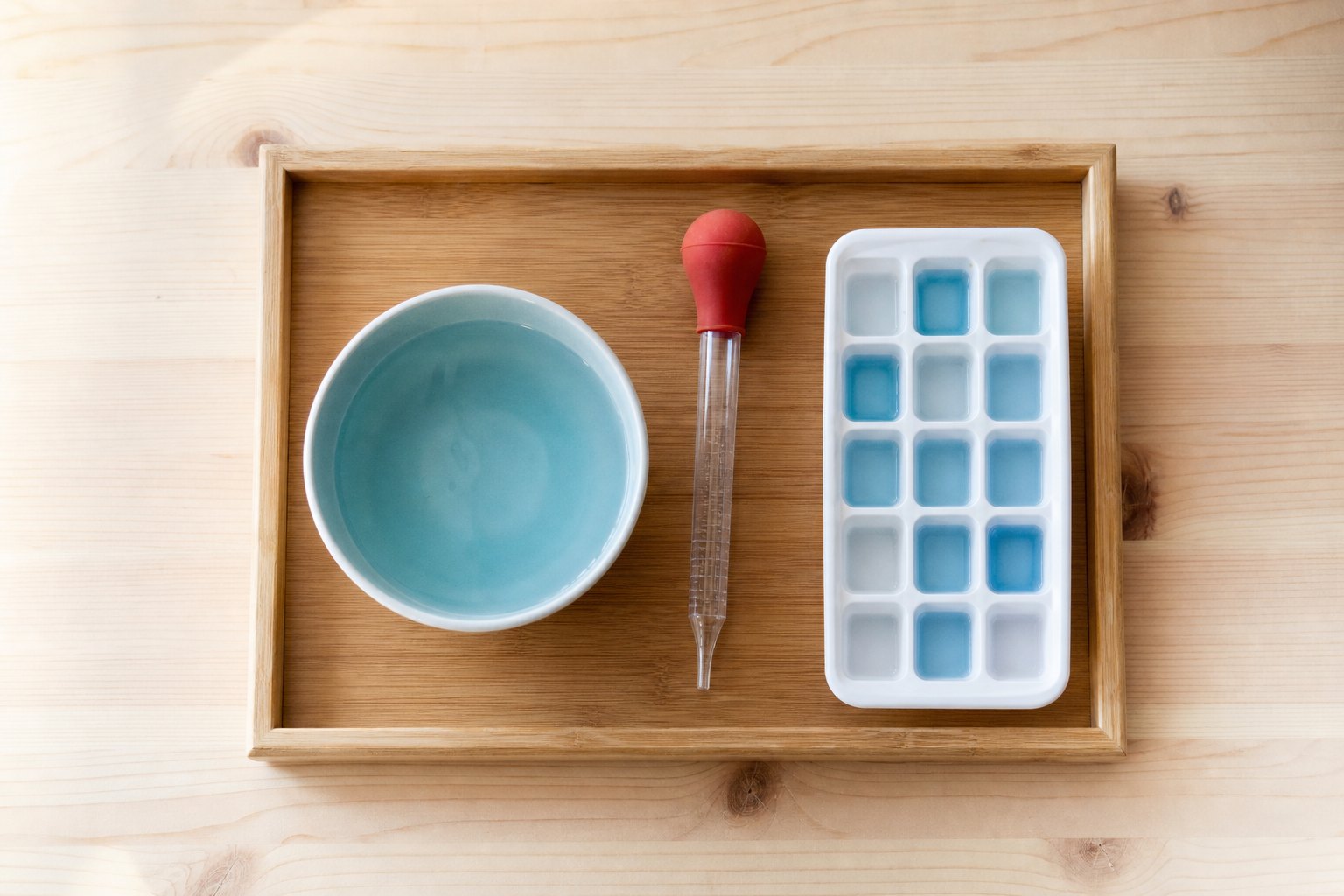 Draufsicht auf ein Holztablett, links eine Keramikschale mit blau eingefaerbtem Wasser, rechts eine weisse Eiswuerfelform mit