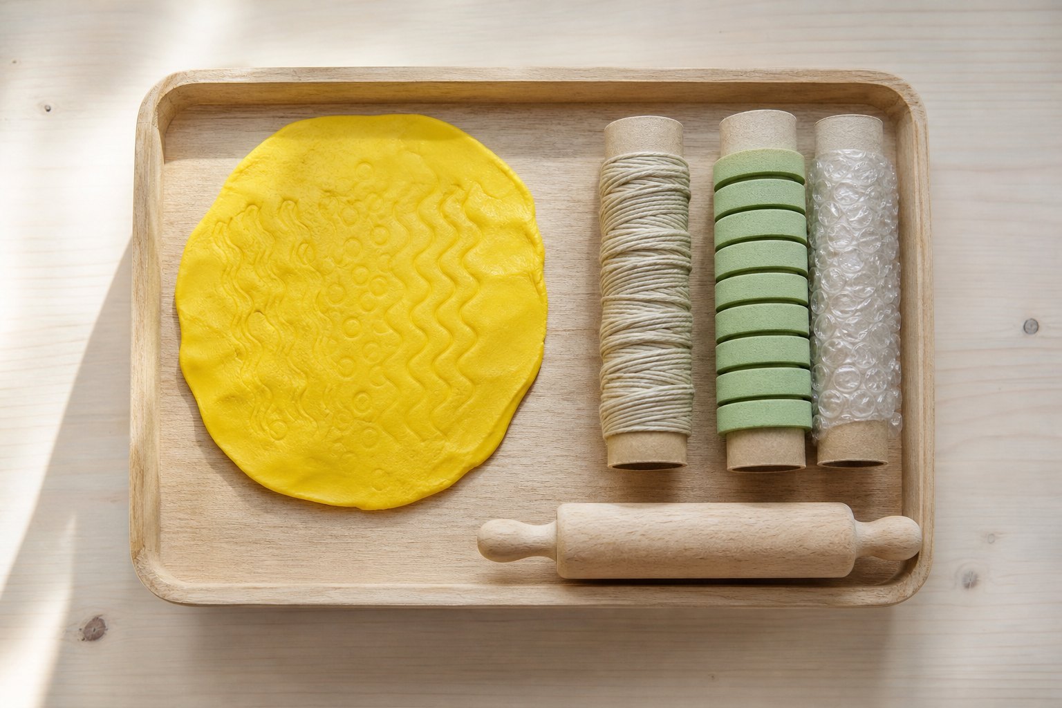 Montessori Tablett mit eine flach ausgerollte Platte gelber Knete mit sichtbaren Rillenmustern, daneben drei Papprollen, eine
