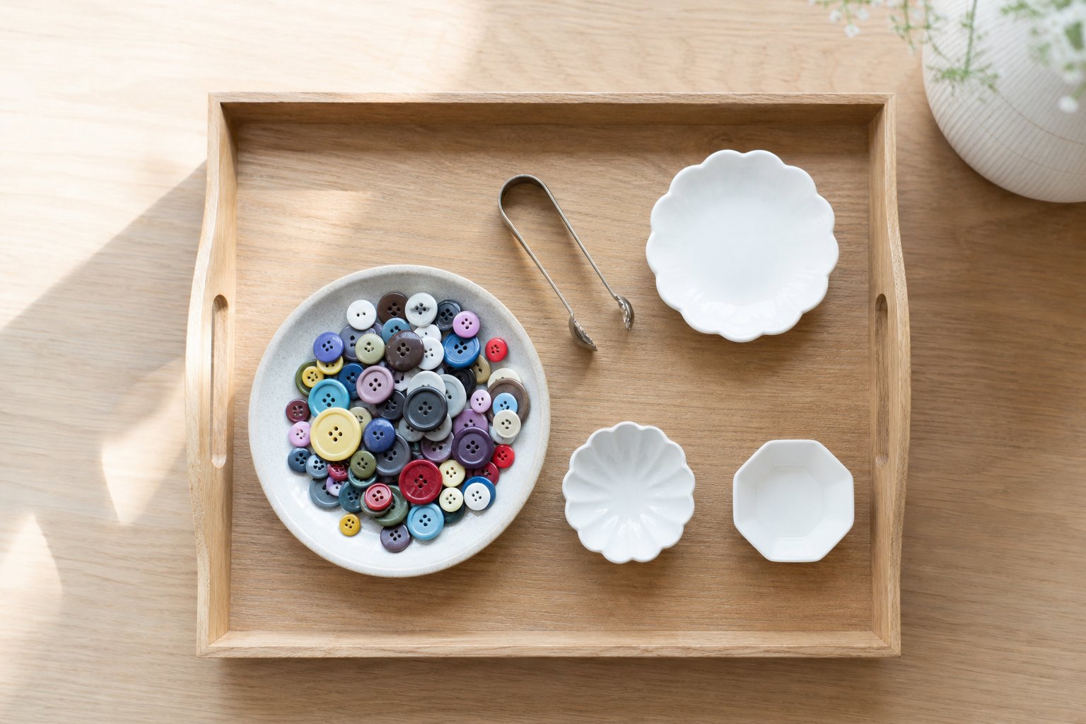 Montessori Tablett mit eine flache Schale mit vielen bunten Knoepfen, drei kleine weiße Schälchen und eine silberne Zuckerzan
