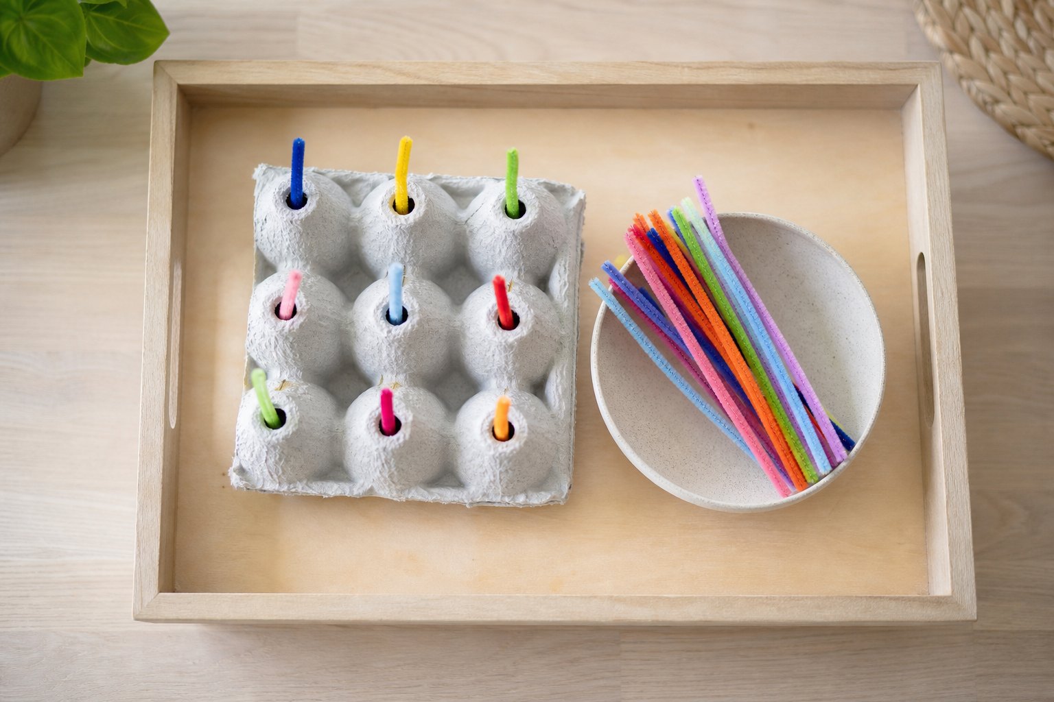 Montessori Tablett mit ein umgedrehter grauer Eierkarton, in dessen Löchern bunte Pfeifenreiniger stecken, daneben weitere Pf