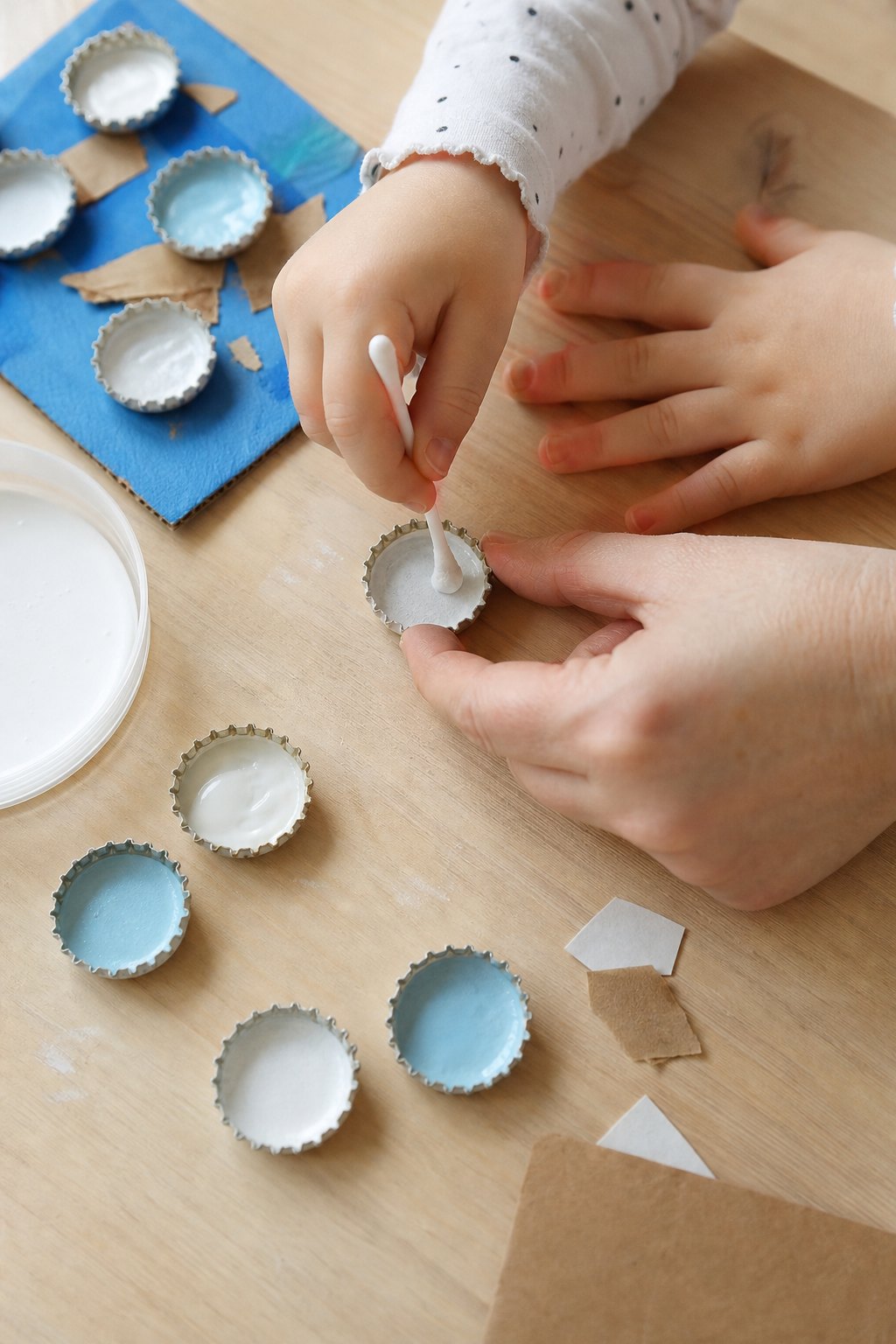 Eine Kinderhand tupft mit dem Wattestäbchen weiße Farbe in einen Kronkorken