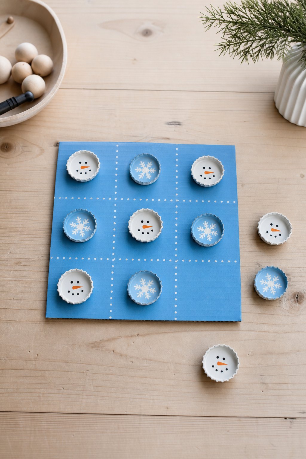 Drei gewinnt mit Schneemännern und Schneeflocken — Ein blau bemaltes Kartonquadrat von 21 cm liegt auf einem hellen Holztisch