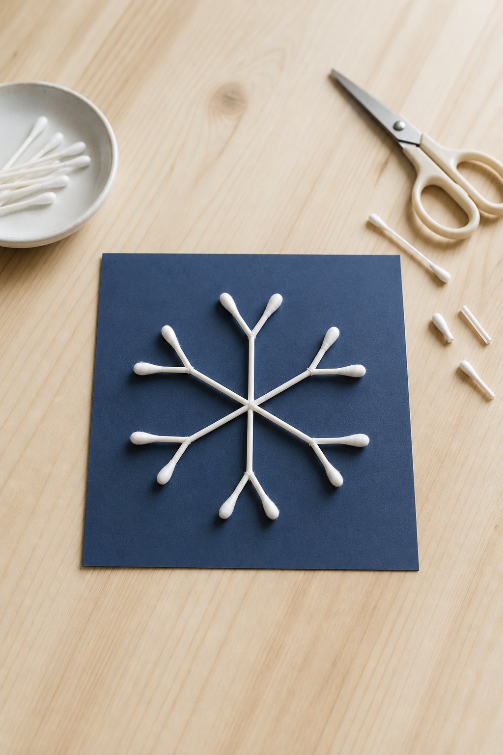 Die Schneeflocke aus Wattestäbchen — Ein dunkelblaues Quadrat aus Tonkarton, 15 cm gross, liegt auf einem hellen Holztisch