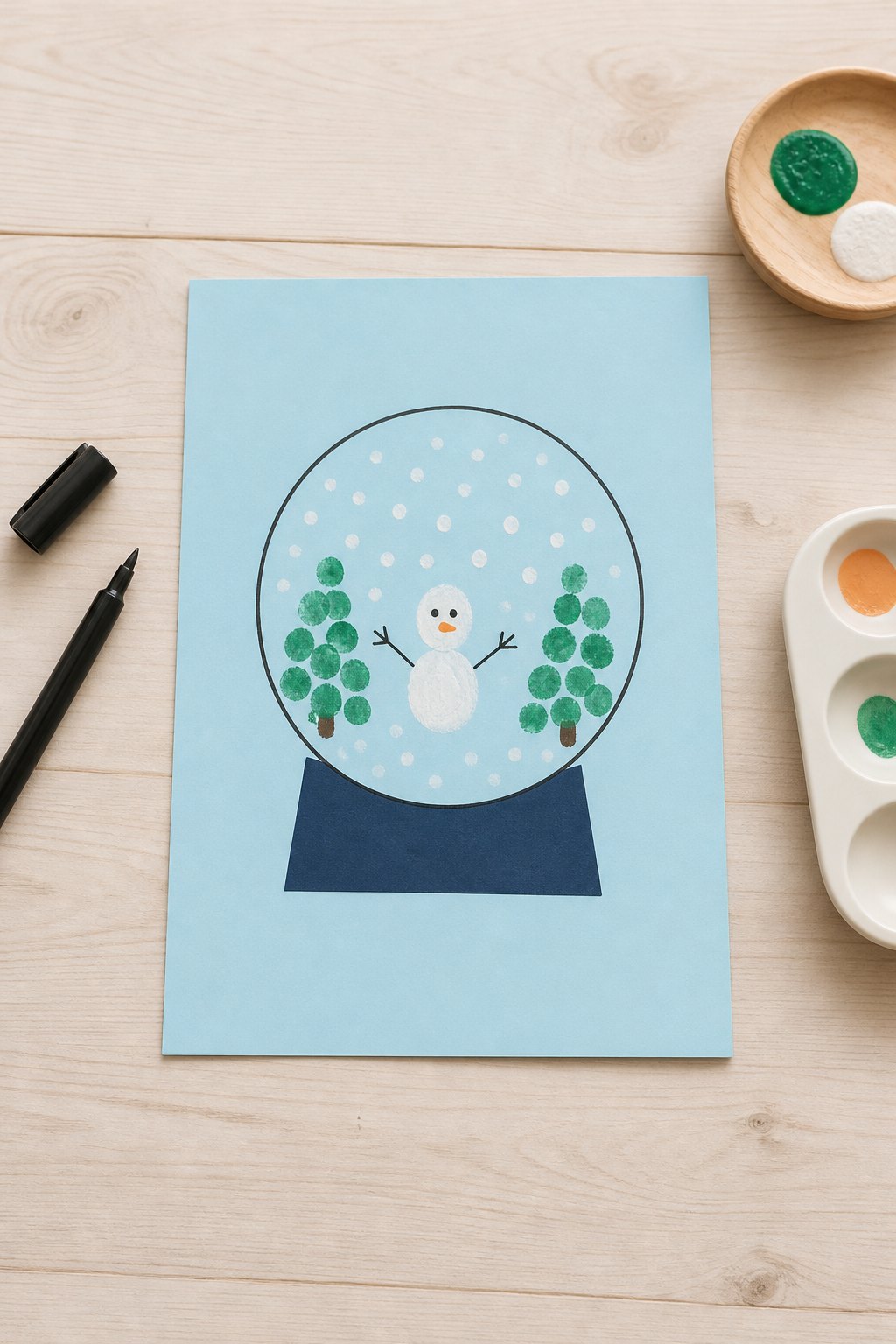 Die gemalte Schneekugel aus Fingerabdrücken — Ein hellblaues Blatt Tonpapier im Format DIN A4 liegt auf einem hellen Holztisc