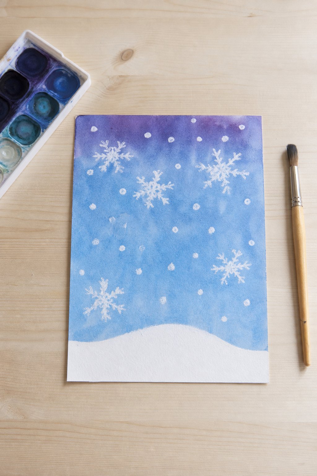 Das Schneebild, das erst mit Farbe erscheint — Ein Blatt Malpapier im Format DIN A4 liegt auf einem hellen Holztisch