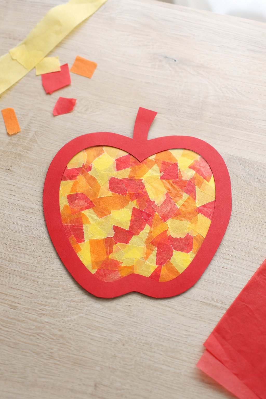 Das Apfel-Fensterbild aus gerissenem Transparentpapier — Ein Fensterbild in Apfelform auf einem hellen Holztisch, von oben fo