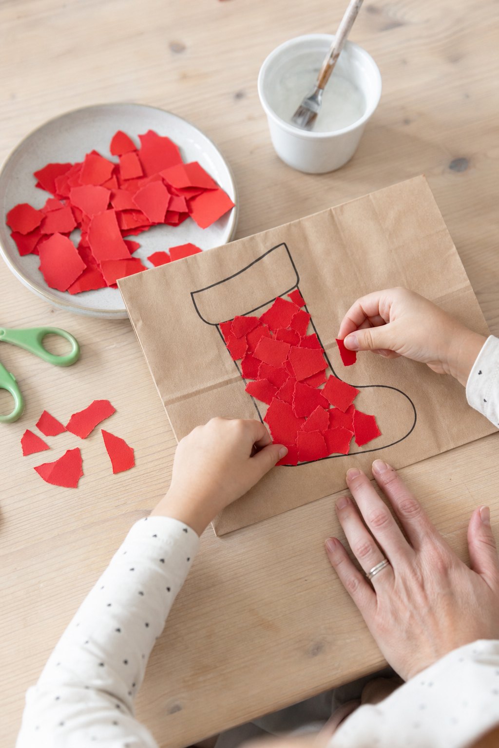 Kinderhände kleben rote Papierschnipsel in eine Stiefelform auf der Papiertüte
